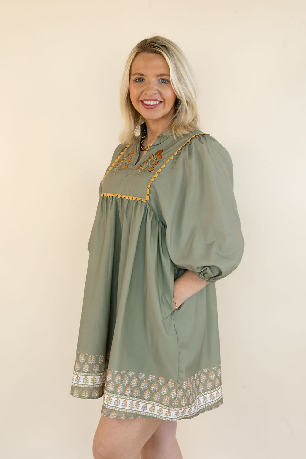 A smiling blonde woman wearing an olive green tunic dress with gold embroidered details and a patterned hem, standing against a plain beige background.