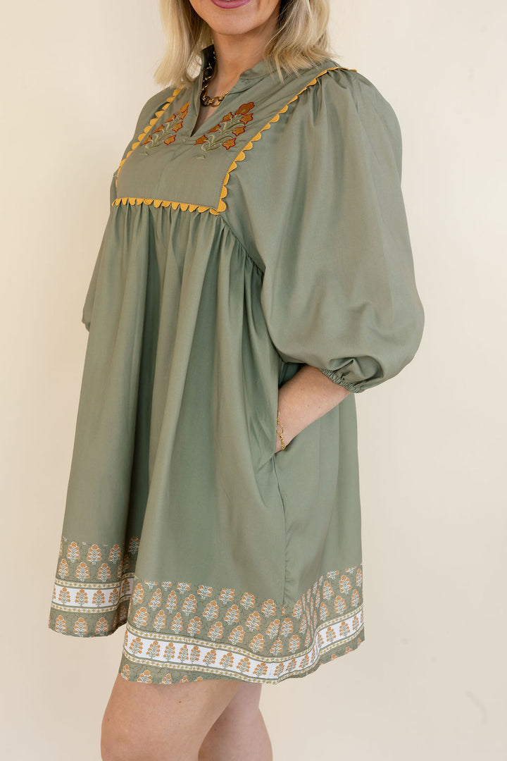 A woman wearing a sage green bohemian-style tunic dress with yellow embroidered details and a patterned hem, photographed against a plain cream background.