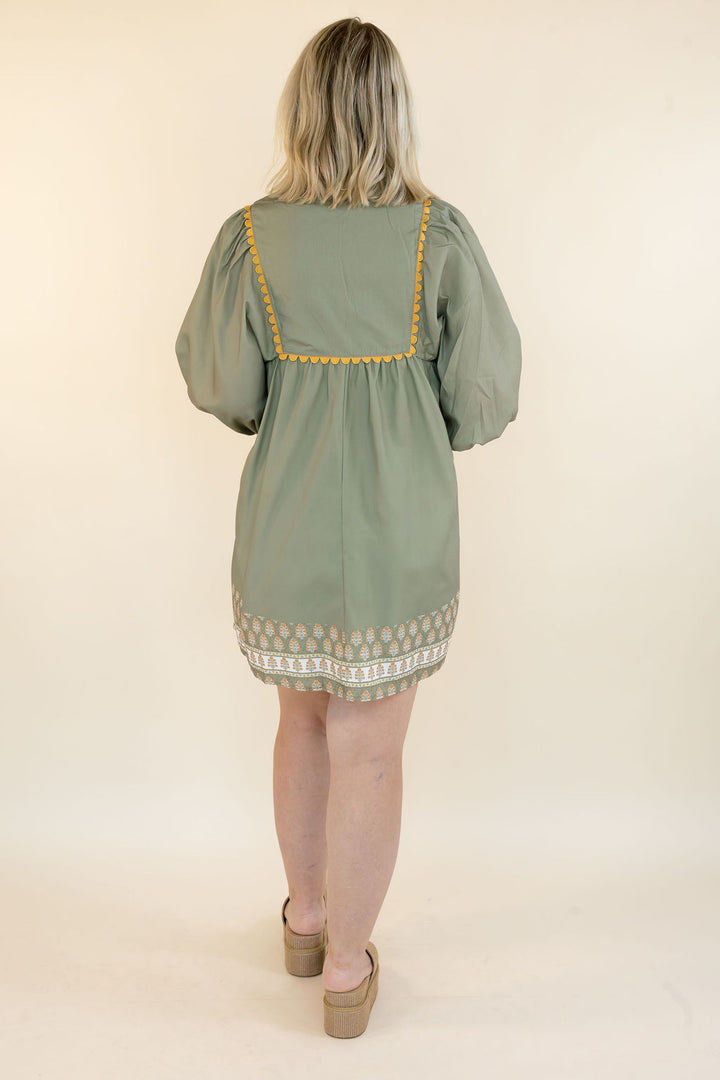 A woman viewed from behind wearing a sage green short dress with yellow embroidered trim and a patterned hem, standing against a plain cream background.