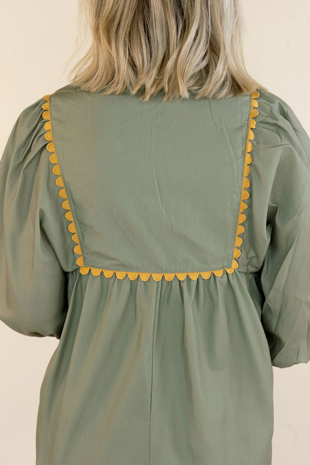 A woman viewed from the back wearing a sage green dress with yellow scalloped trim along the neckline and waist.