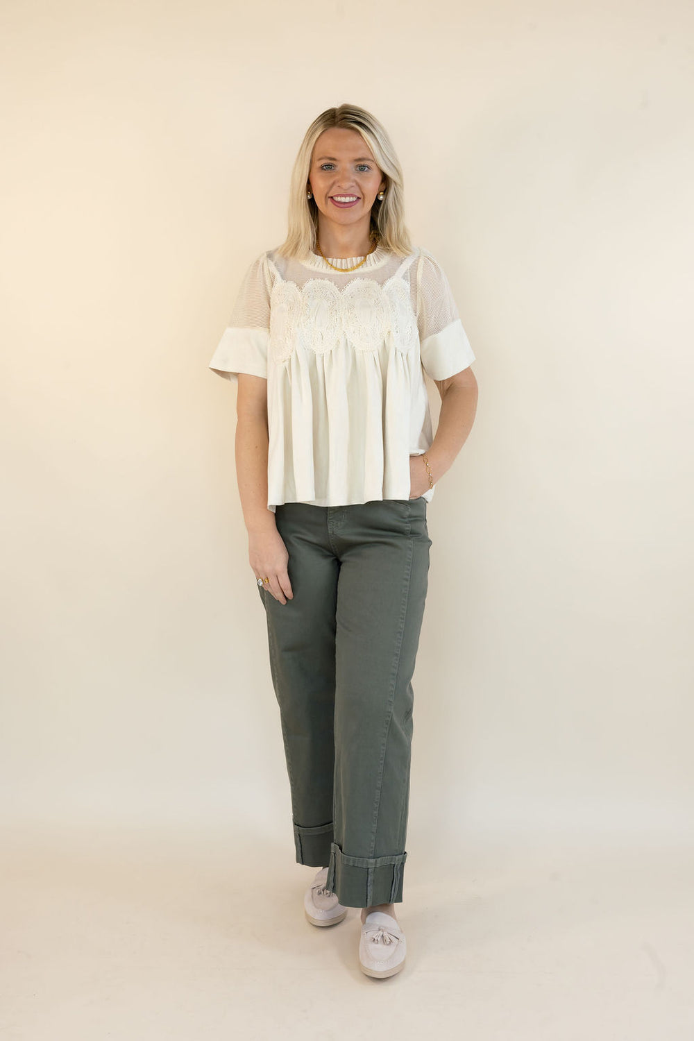 A woman modeling a white short-sleeve blouse with a sheer yoke and pleated front, paired with olive green pants and white sneakers, standing against a plain cream studio background.
