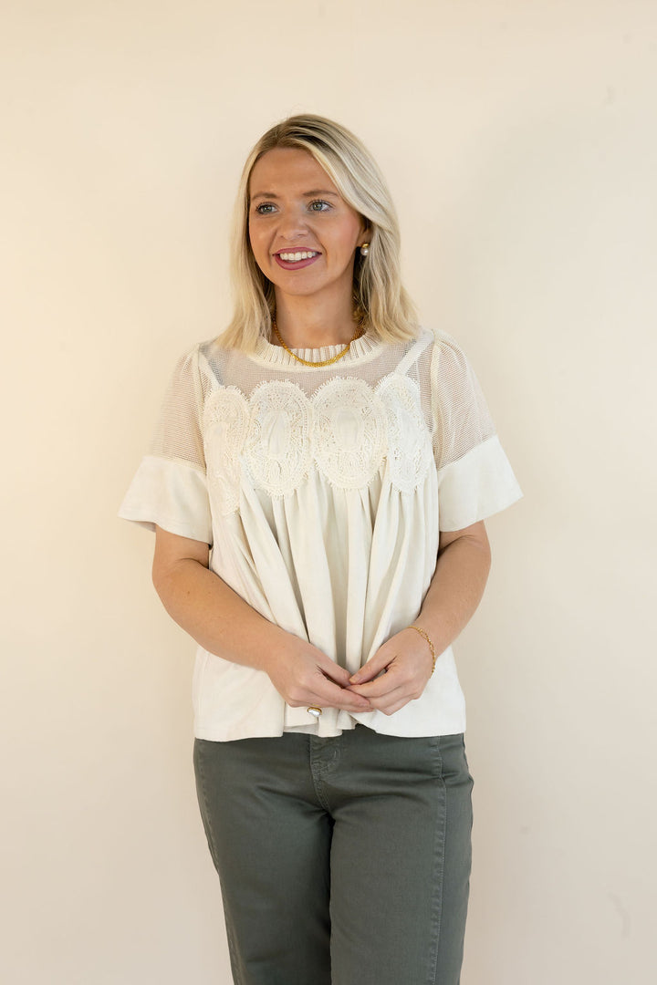 A woman with blonde hair wearing a cream-colored blouse with sheer sleeves and a floral lace bodice, paired with dark gray pants, standing against a plain beige wall.
