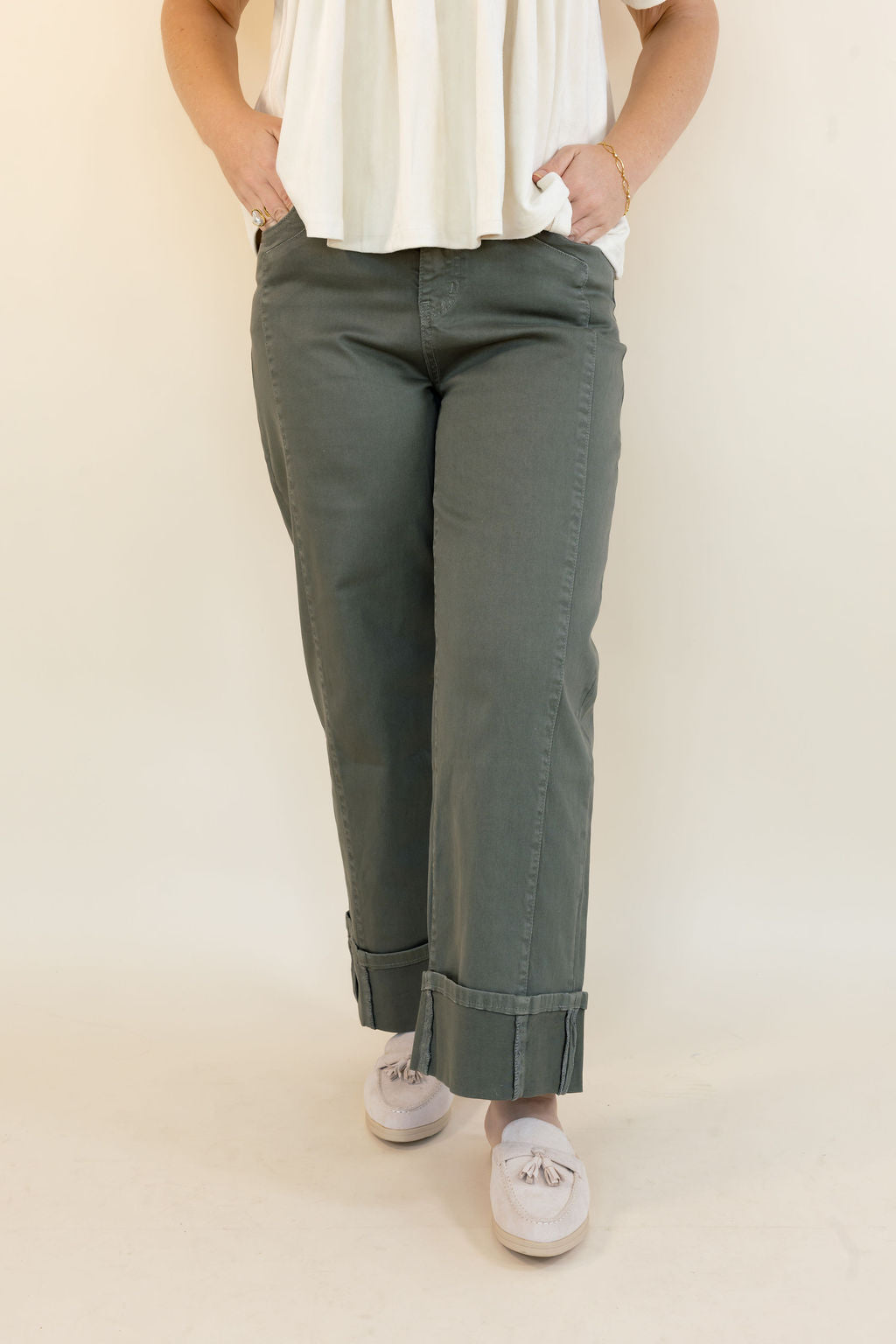 A person wearing olive green cropped pants with rolled cuffs, paired with a white top and white sneakers, standing against a plain cream background.