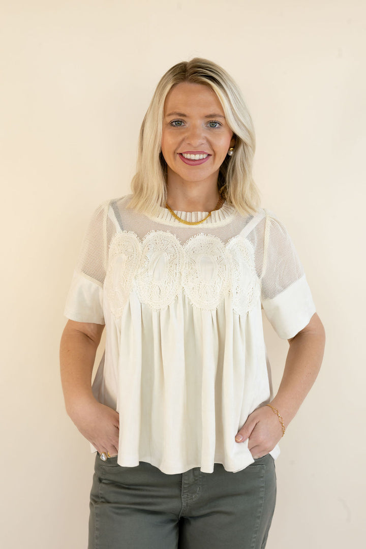 A smiling blonde woman wearing a cream-colored short-sleeve blouse with a sheer mesh yoke and intricate lace applique across the chest, paired with dark gray pants, standing against a plain beige background.