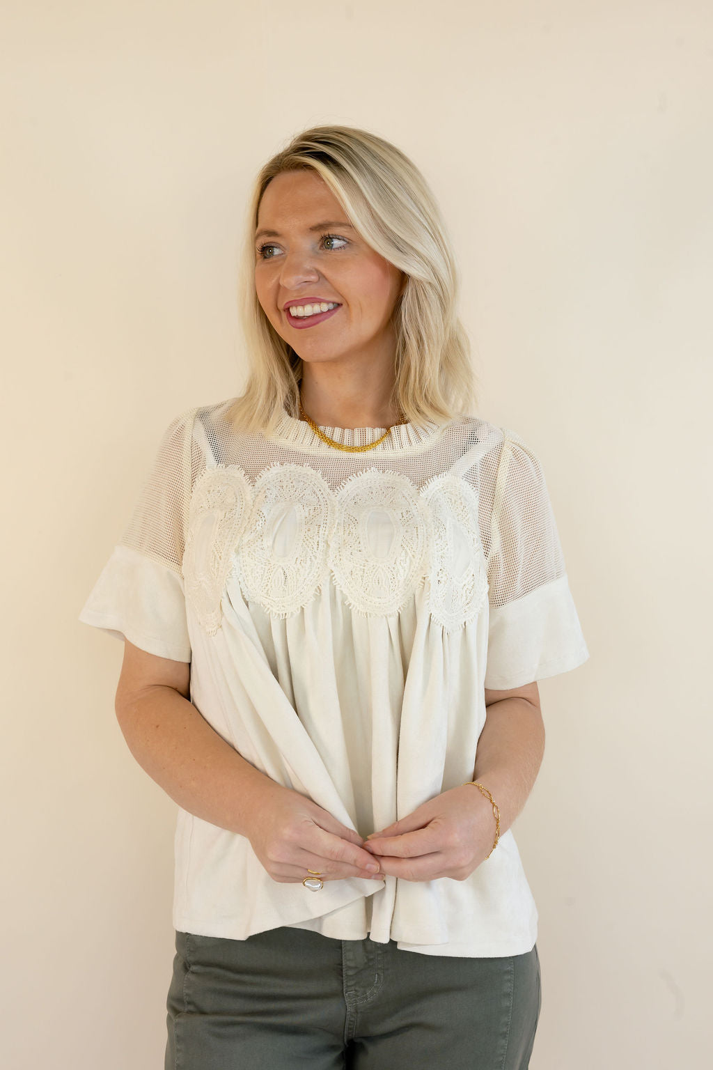 A woman with blonde hair wearing a white short-sleeve blouse with lace applique and grey pants, standing against a plain beige wall.
