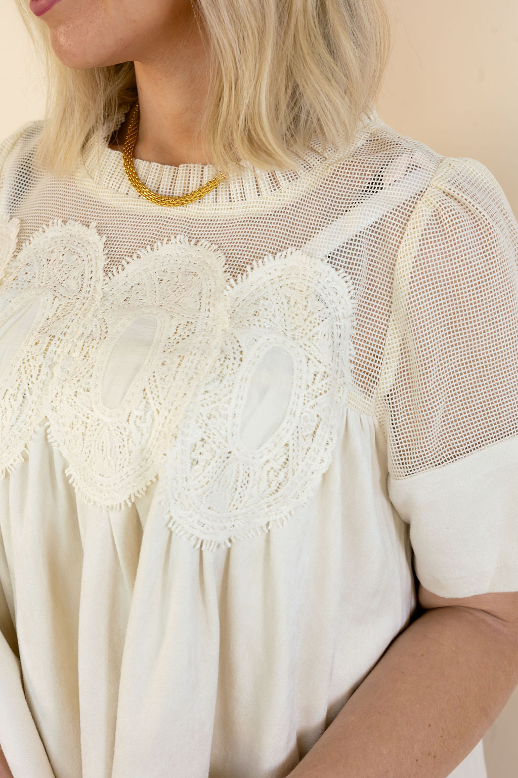 A close-up of a woman wearing a cream-colored blouse with sheer mesh sleeves, a high gathered neckline, and a large scalloped lace yoke across the chest.