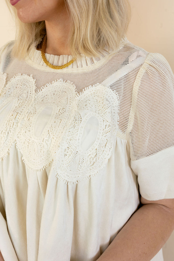 A close-up of a woman wearing a cream-colored blouse with sheer mesh sleeves, a high gathered neckline, and a large scalloped lace yoke across the chest.