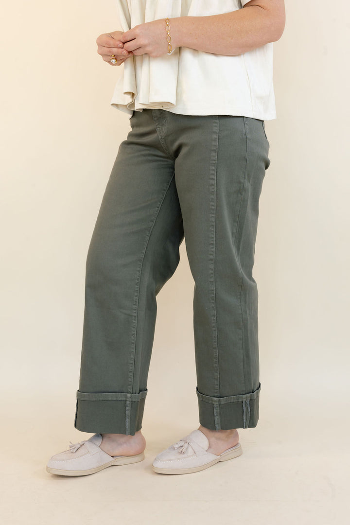 A person wearing olive green cuffed pants, a cream blouse, and off-white slip-on shoes, standing against a plain beige studio background.