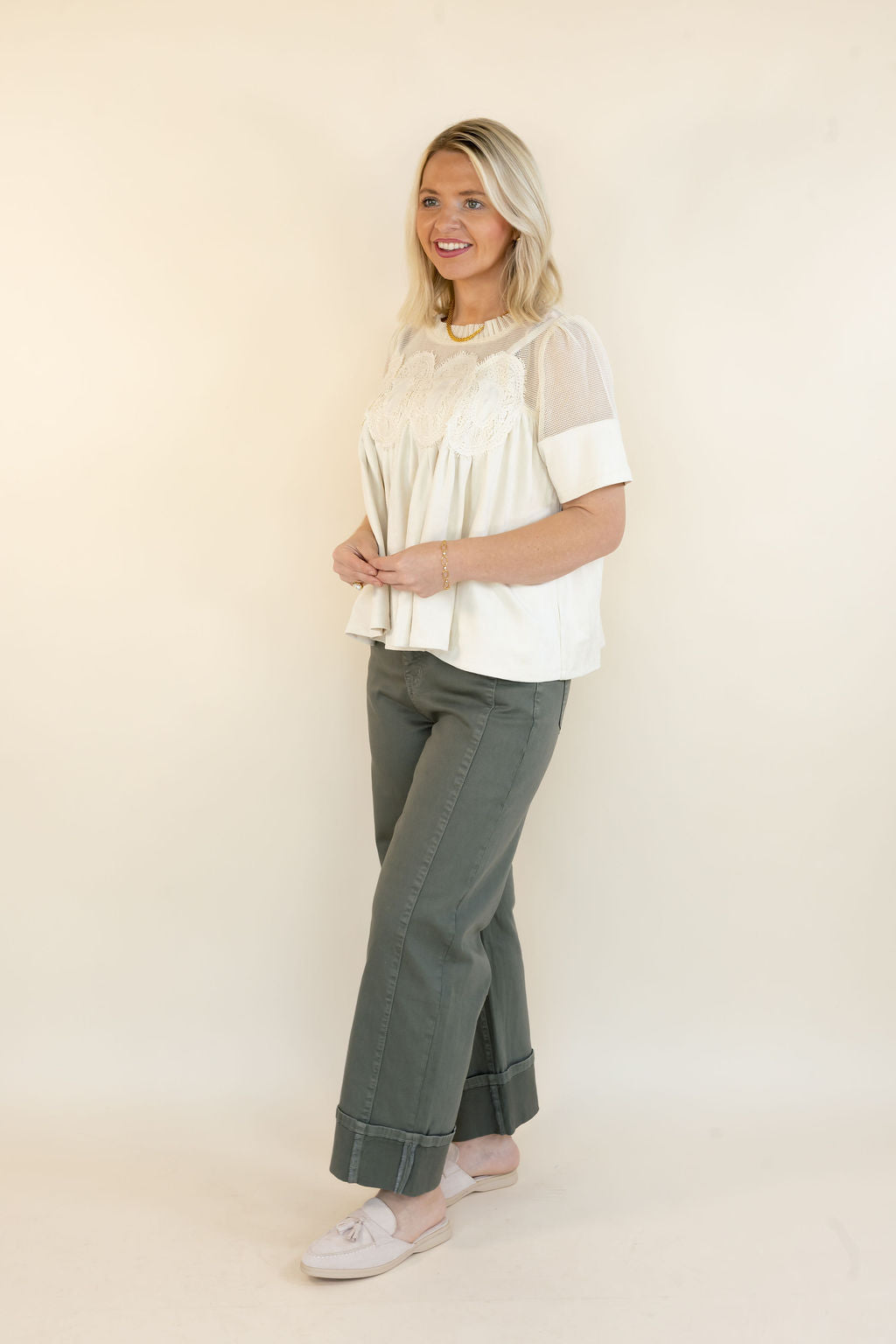 A woman modeling a cream short-sleeve blouse with sheer sleeves and a lace yoke, paired with olive green wide-leg pants and white slip-on shoes, against a plain cream studio background.