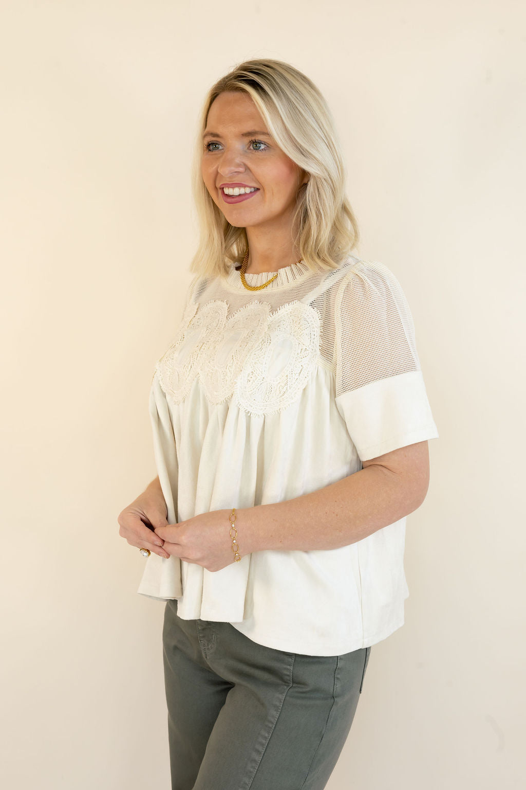 A woman modeling a cream-colored short-sleeve blouse with sheer mesh sleeves and a floral lace yoke, paired with olive green pants, against a plain beige background.
