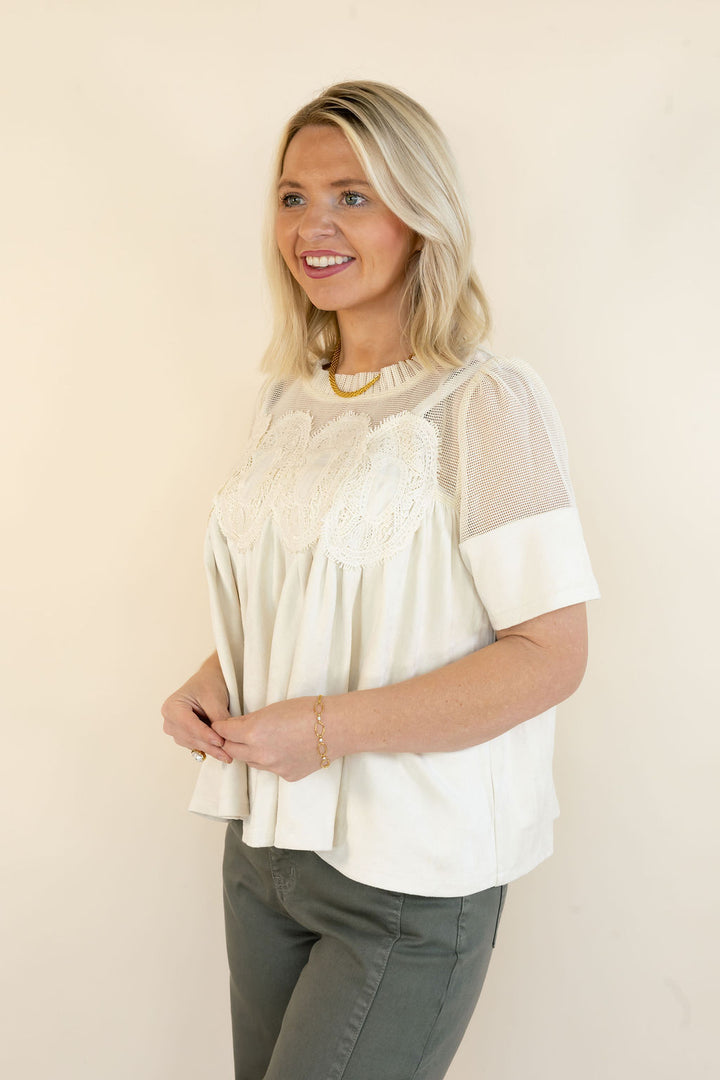 A woman modeling a cream-colored short-sleeve blouse with sheer mesh sleeves and a floral lace yoke, paired with olive green pants, against a plain beige background.