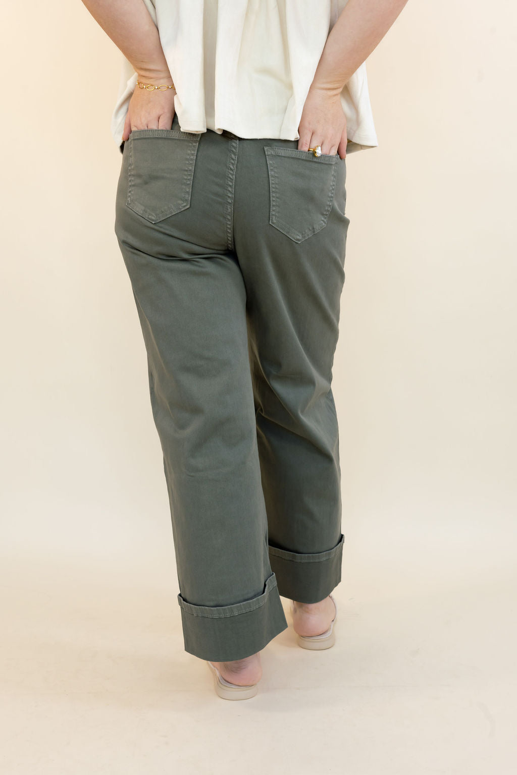 A person shown from the back wearing olive green wide-leg cropped pants with rolled cuffs and a cream-colored top, standing against a plain beige studio background.