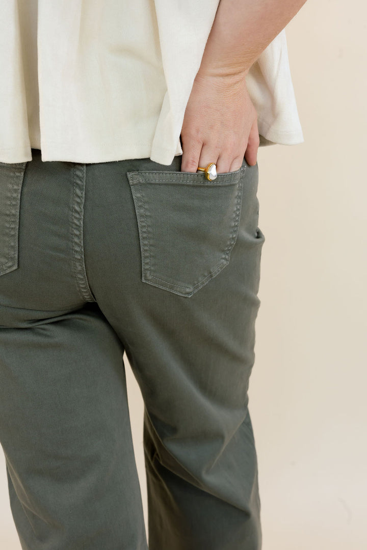 Close-up of a person wearing dark olive green pants, showing the back pocket and hand in a cream-colored top against a plain beige background.