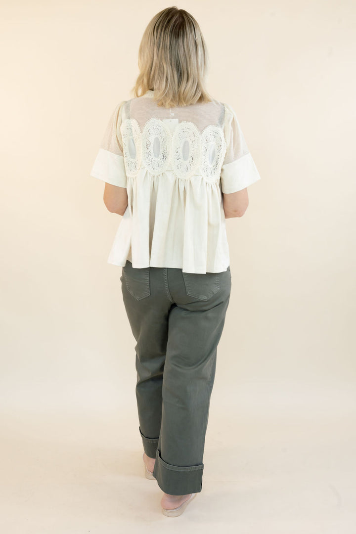 A woman viewed from behind wearing a cream-colored blouse with a sheer lace back panel and short sleeves, paired with dark gray pants, against a plain beige studio background.