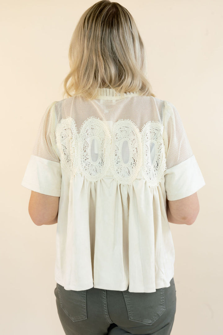 A woman viewed from behind wearing a short-sleeve cream blouse with sheer mesh and intricate lace detailing across the upper back, paired with dark gray pants, against a solid light beige background.