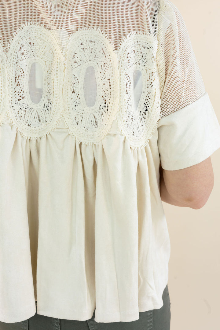 Close-up of a cream-colored blouse with a sheer mesh back panel, three large scalloped lace appliqués, and rolled short sleeves, worn by a model.