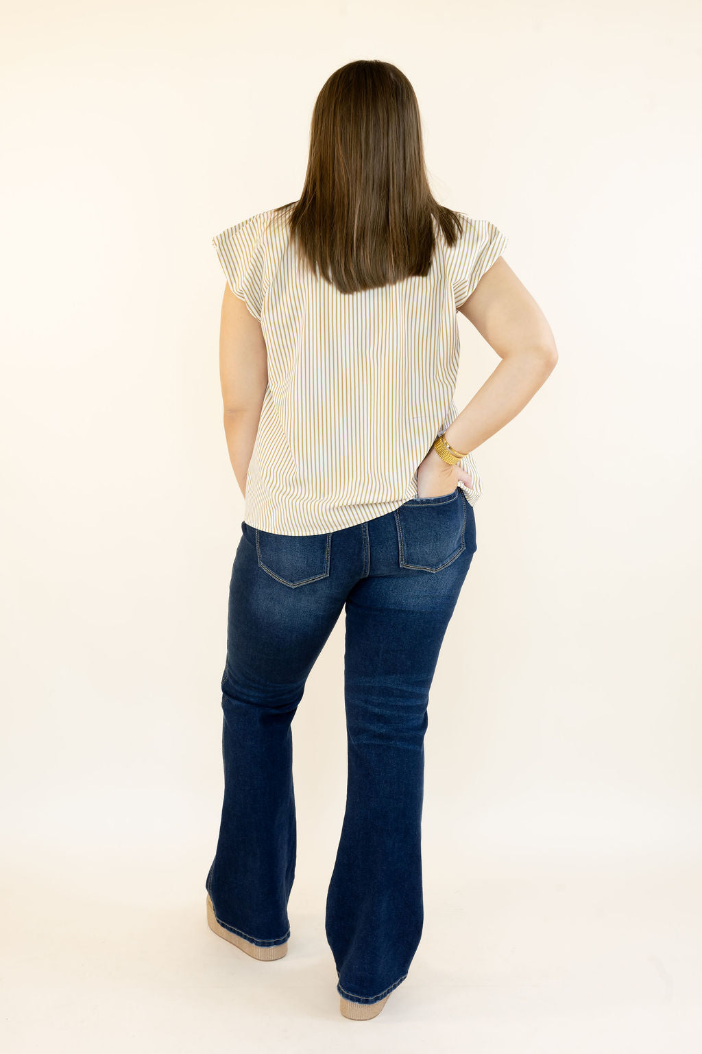 A woman viewed from behind wearing a cream and navy vertically striped short-sleeve top and dark blue flared jeans, standing against a plain off-white studio background.