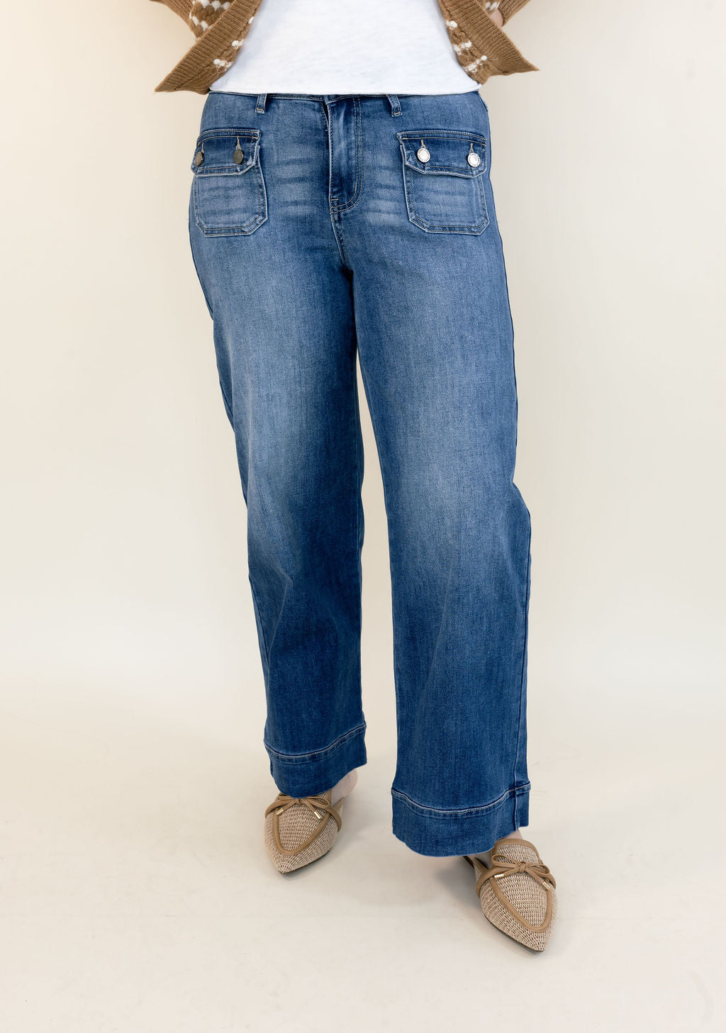 A person wearing blue wide-leg jeans with a faded wash, styled with a white top and beige pointed-toe flats against a plain light beige studio background.