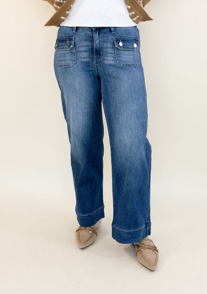 A person wearing blue wide-leg jeans with a faded wash, styled with a white top and beige pointed-toe flats against a plain light beige studio background.