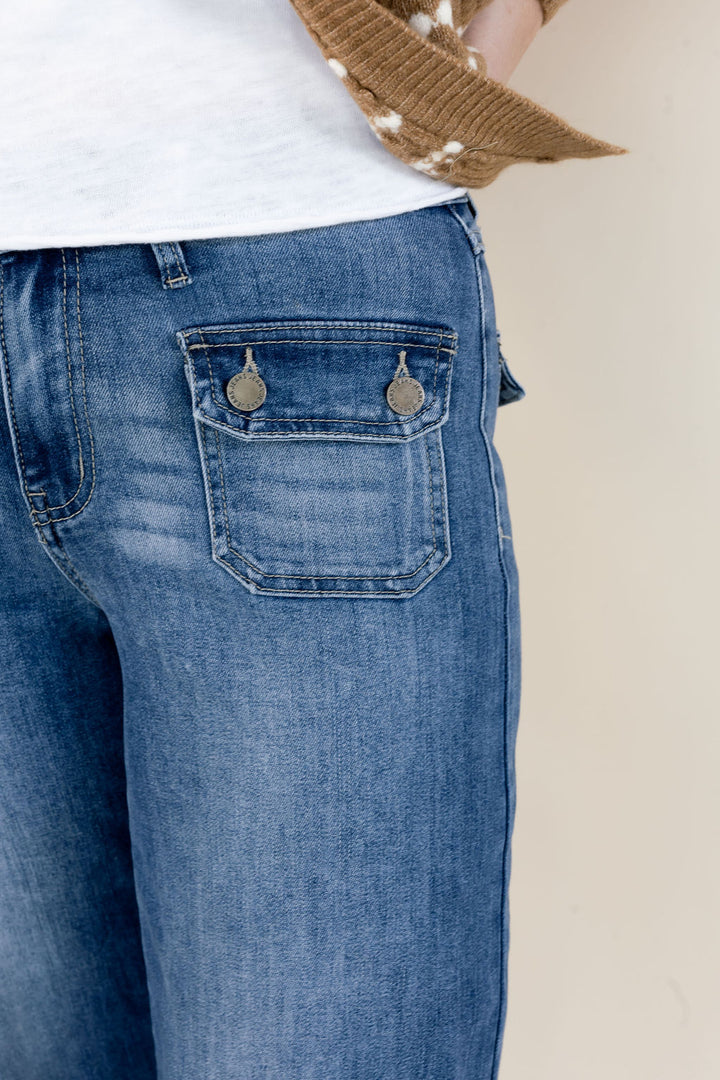 Close-up of a person wearing medium-wash blue denim jeans, showing the back pocket with two metal button details, a white top, and a brown knit hat against a light beige background.