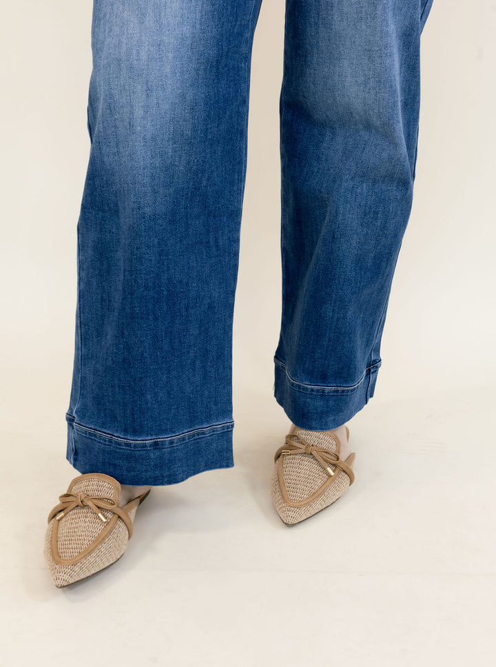 A person wearing blue wide-leg jeans and beige woven pointed-toe flats with bow details, shown from the knees down against a plain light background.