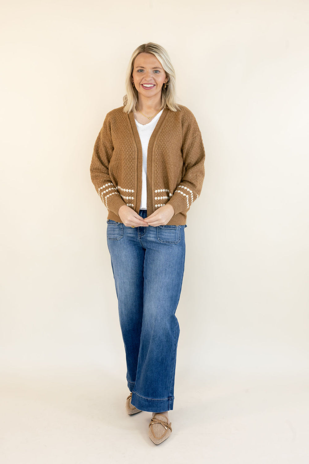 A woman stands against a plain cream studio background wearing a brown knit cardigan with cream knit sleeve trim, a white top, blue jeans, and tan pointed flats.