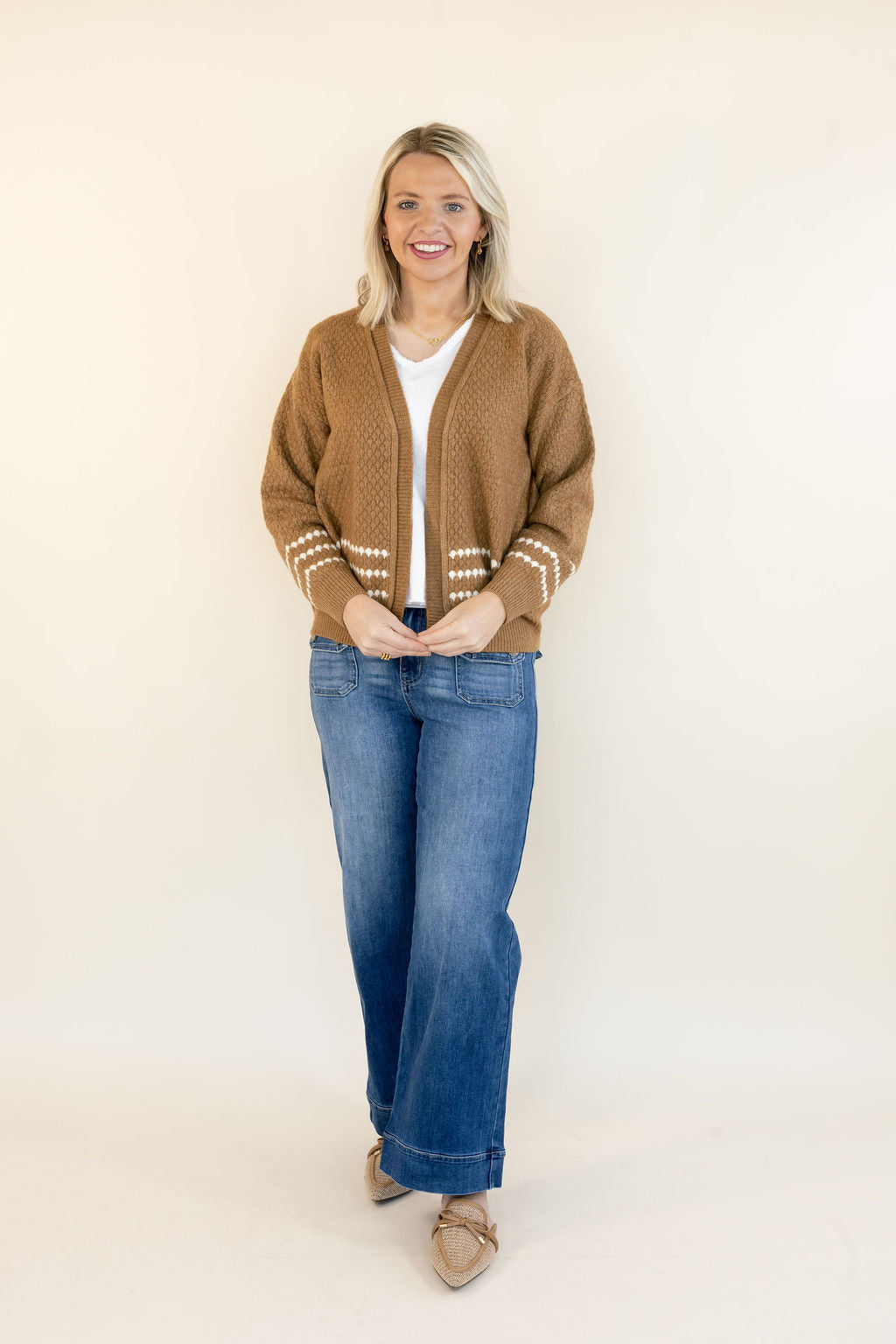 A woman stands against a plain cream studio background wearing a brown knit cardigan with cream knit sleeve trim, a white top, blue jeans, and tan pointed flats.