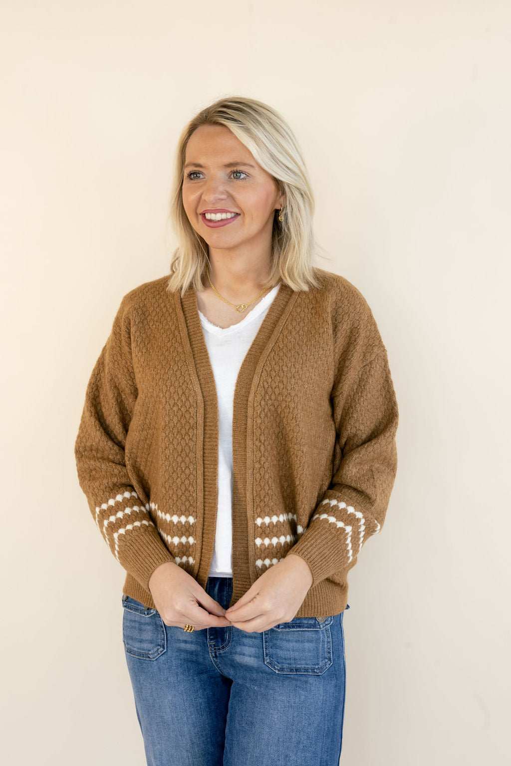 A woman with blonde hair wearing a brown cable-knit cardigan with white geometric patterns on the cuffs and hem, styled over a white t-shirt and blue jeans, standing against a plain beige background.