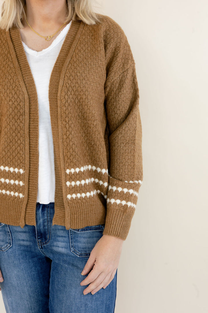 A woman wearing a brown cable-knit cardigan with white geometric patterns on the cuffs and hem, styled over a white t-shirt and blue jeans.