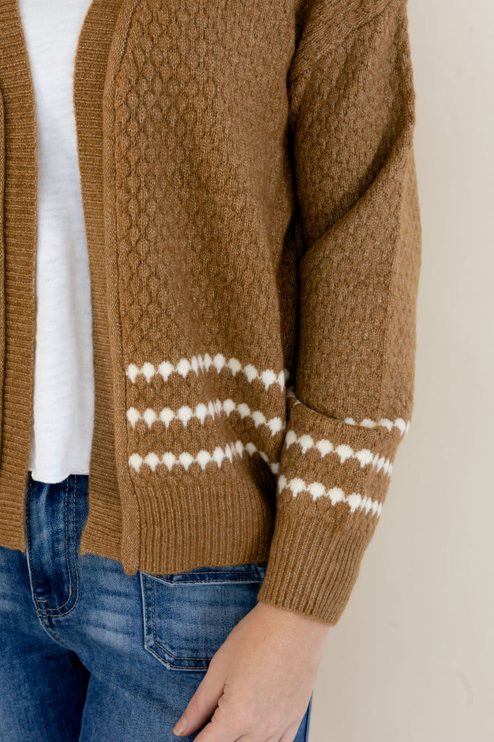 A close-up of a person wearing a brown cable-knit cardigan with white patterned stripes near the cuffs, layered over a white top and blue jeans.