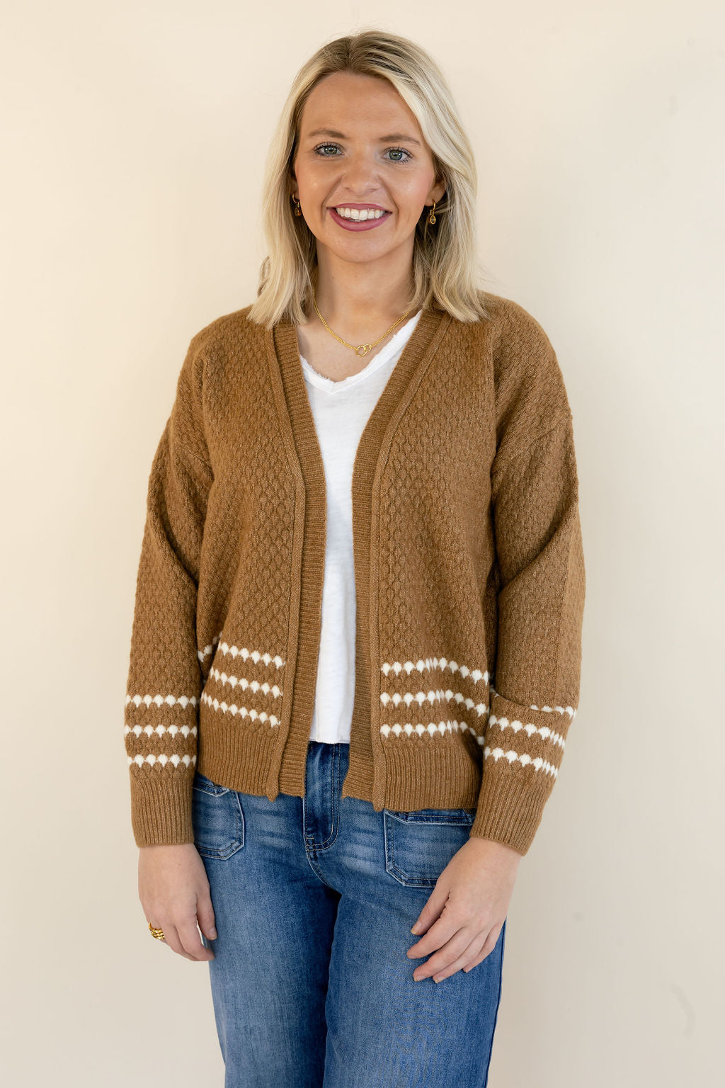 A smiling blonde woman wearing a brown cable-knit cardigan with white geometric patterns on the cuffs and hem, styled over a white t-shirt and blue jeans, standing against a plain beige wall.