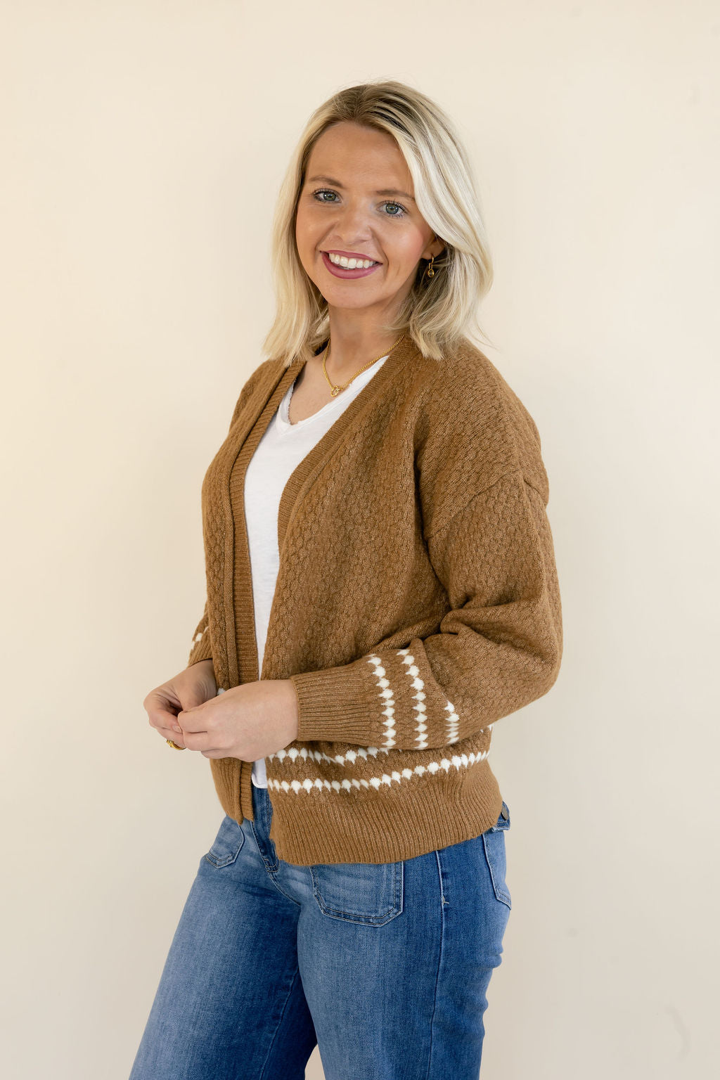 A smiling blonde woman wearing a brown knit cardigan with cream-colored horizontal stripes near the cuffs and hem, styled over a white top and blue jeans, standing against a plain beige background.