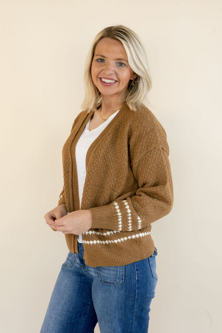 A smiling blonde woman wearing a brown knit cardigan with cream-colored horizontal stripes near the cuffs and hem, styled over a white top and blue jeans, standing against a plain beige background.