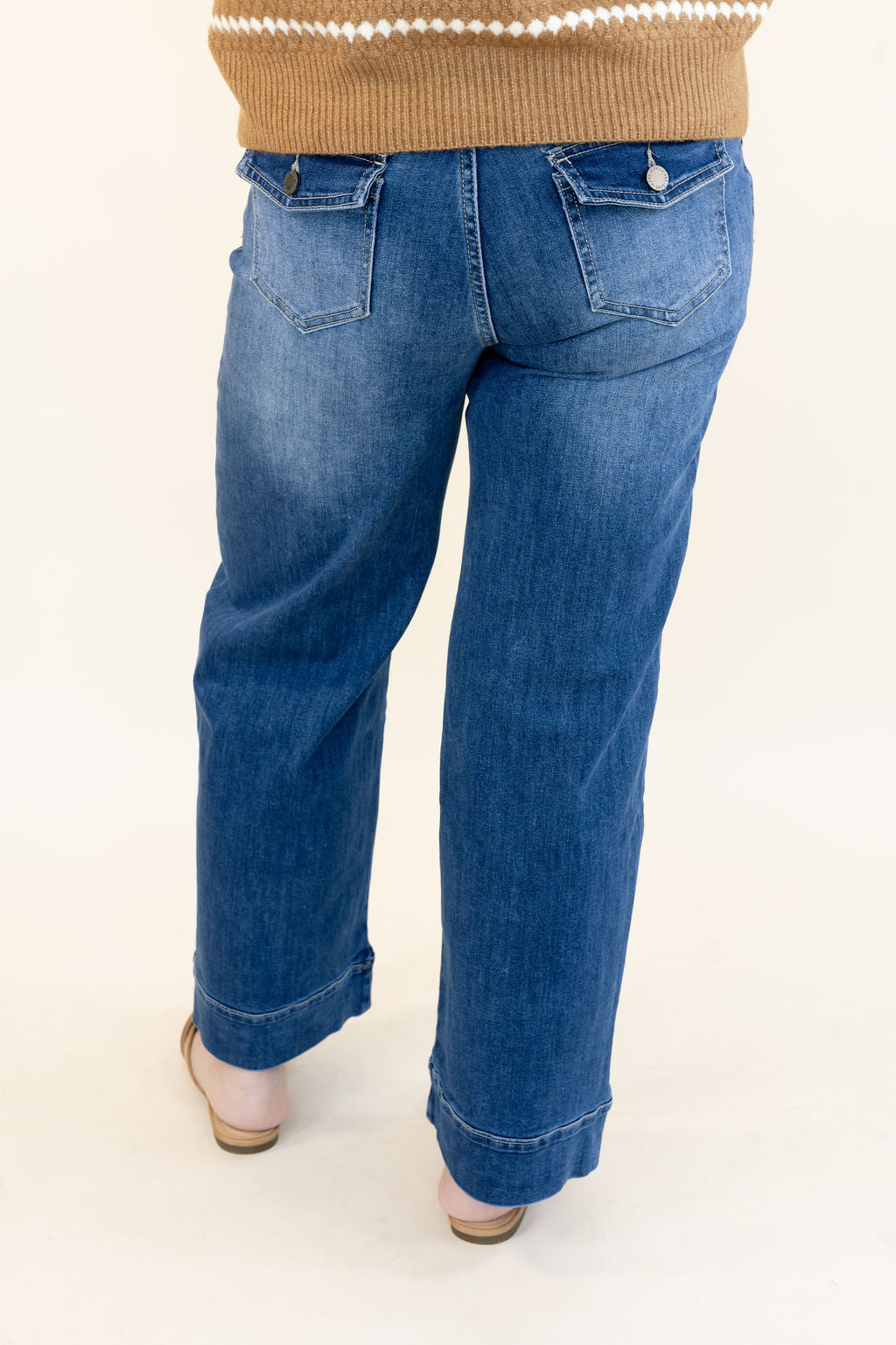 A person shown from the back wearing medium-wash blue jeans with a tan sweater and beige heels, photographed against a plain light background.
