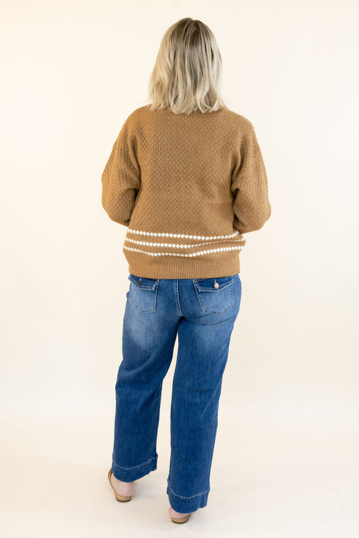 A woman viewed from behind wearing a tan knit sweater with white horizontal stripes near the hem, paired with blue jeans and beige shoes, against a plain off-white studio background.