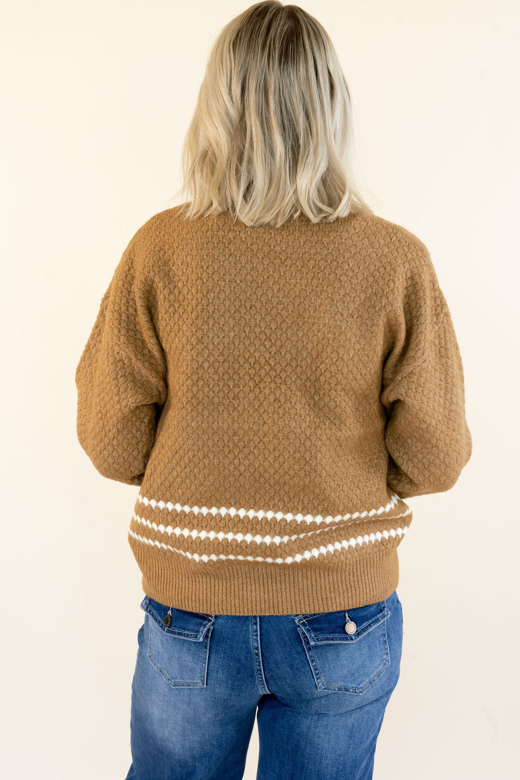A woman viewed from behind wearing a textured tan knit sweater with white geometric patterns along the hem, paired with blue jeans, against a plain off-white background.