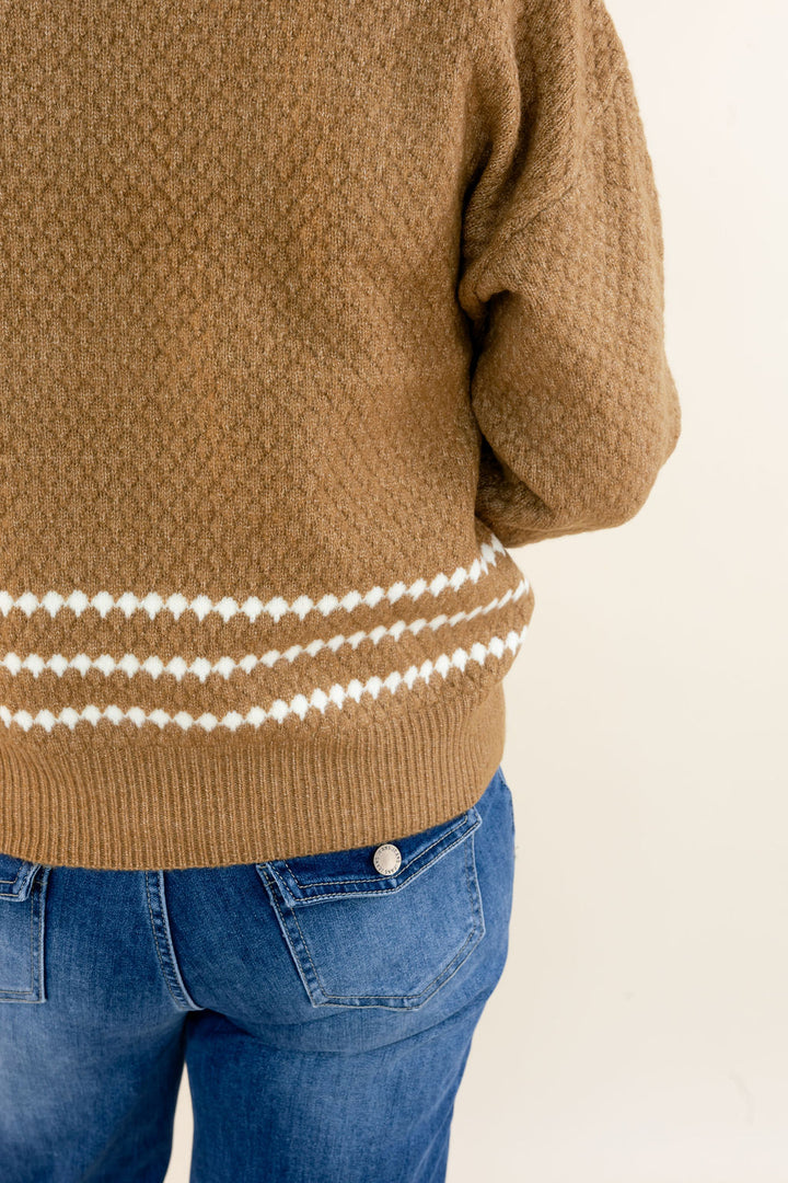 A close-up of a person wearing a textured brown knit sweater with horizontal white diamond-patterned stripes near the hem, paired with blue jeans against a plain light background.