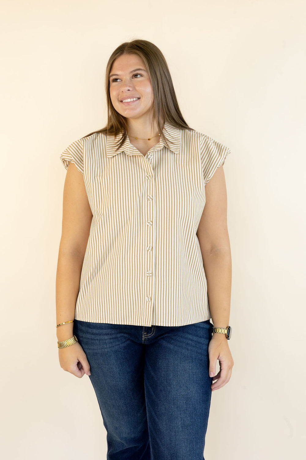 A woman modeling a sleeveless button-up shirt with vertical beige and white stripes, paired with dark blue jeans, standing against a plain off-white background.