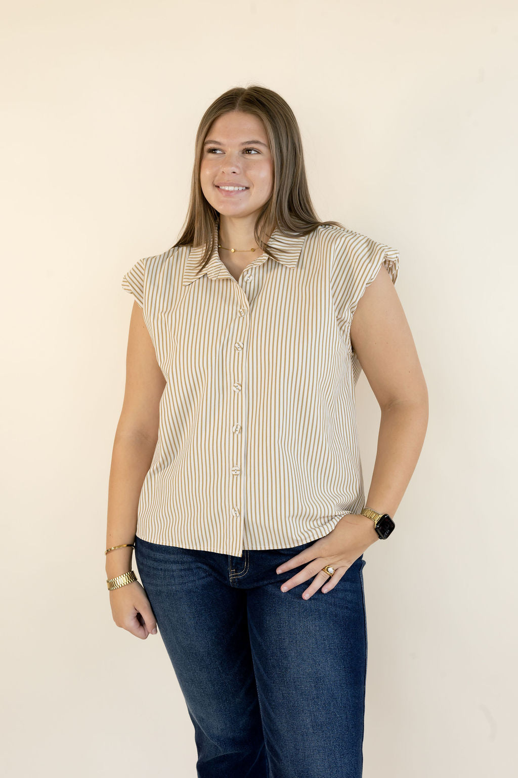A woman modeling a sleeveless button-up shirt with vertical beige and white stripes, paired with dark blue jeans, standing against a plain off-white background.