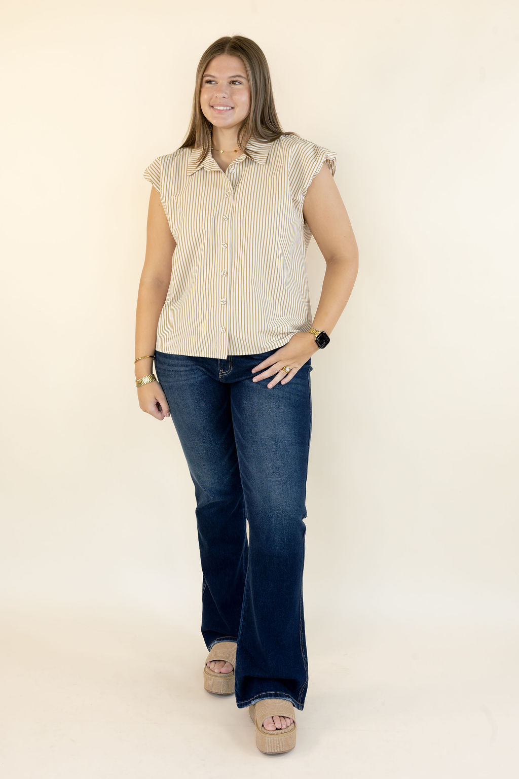 A woman modeling a sleeveless beige and white vertically striped button-down shirt with ruffled cap sleeves, paired with dark blue jeans and beige platform sandals, standing against a plain off-white studio background.