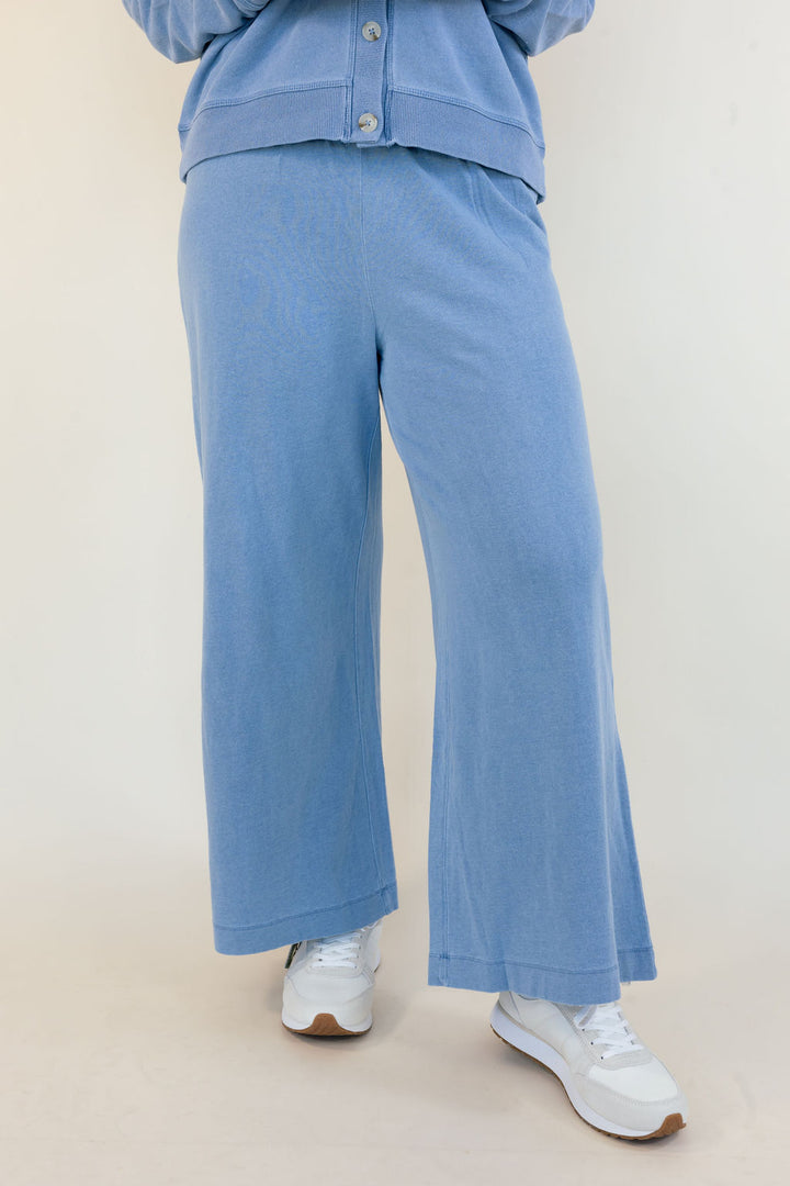 A person wearing light blue wide-leg pants and a matching light blue cropped button-front top, paired with white sneakers, photographed against a plain light beige studio background.
