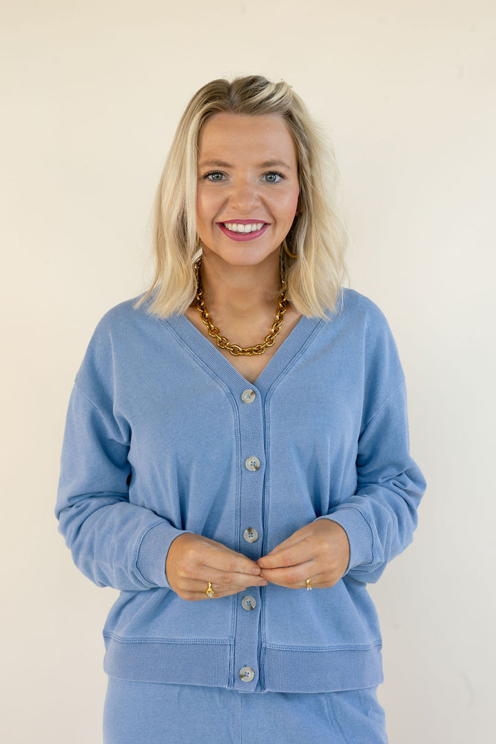 A smiling blonde woman wearing a light blue button-up cardigan and matching bottoms, accessorized with a chunky gold chain necklace, standing against a plain off-white background.