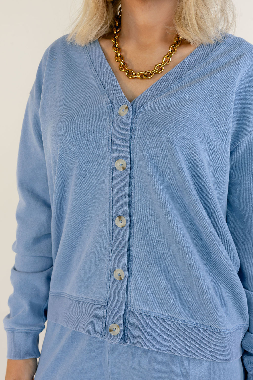 A close-up of a person wearing a light blue long-sleeve button-front cardigan with ribbed trim, paired with a chunky gold chain necklace against a plain light background.