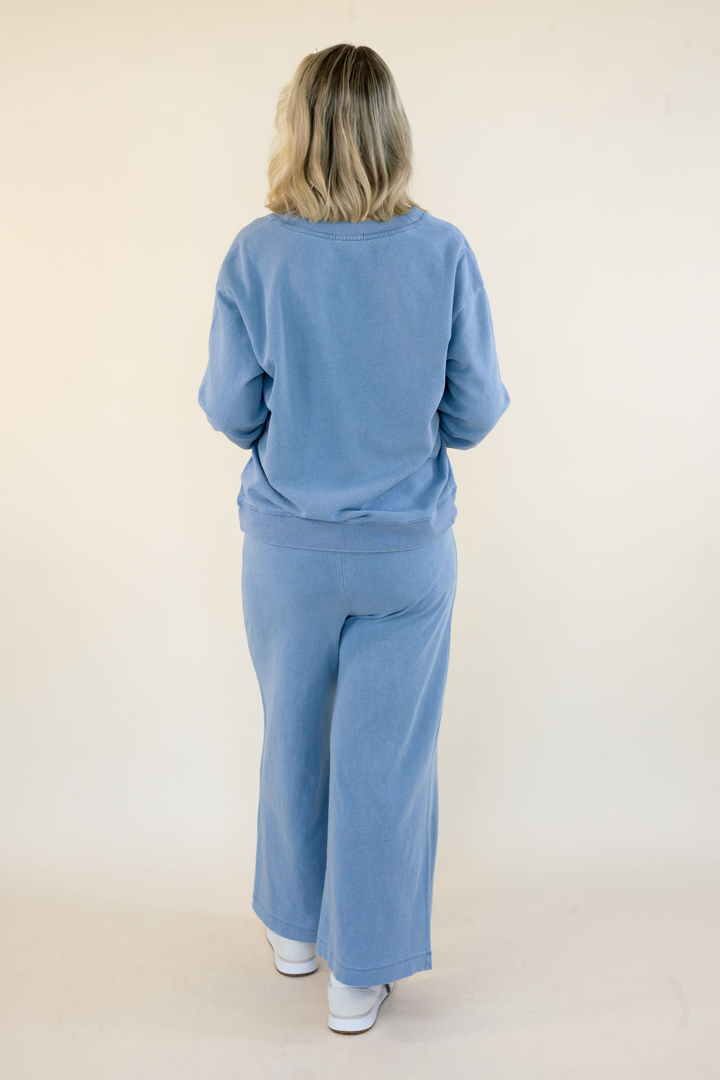 A woman viewed from behind wearing a matching light blue sweatshirt and wide-leg pants set, standing against a plain cream studio background.