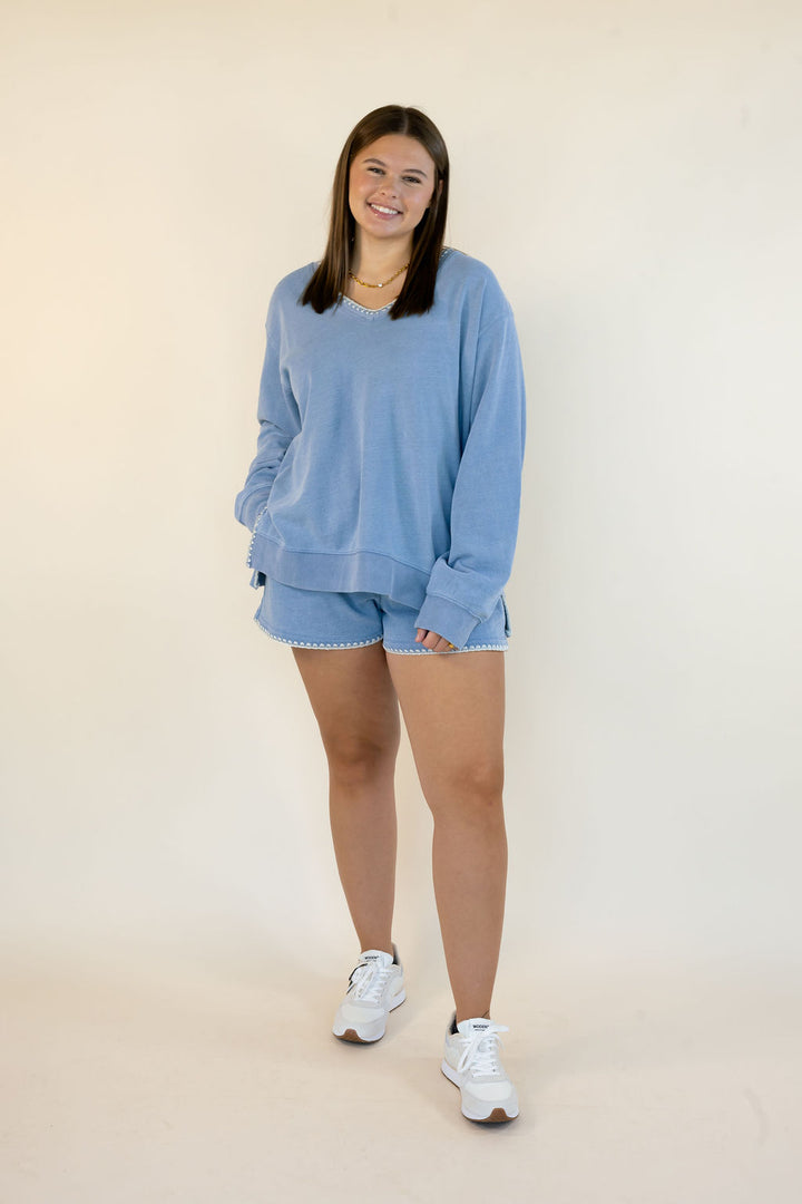 A woman modeling a light blue sweatshirt and matching shorts set, paired with white sneakers, standing against a plain off-white studio background.