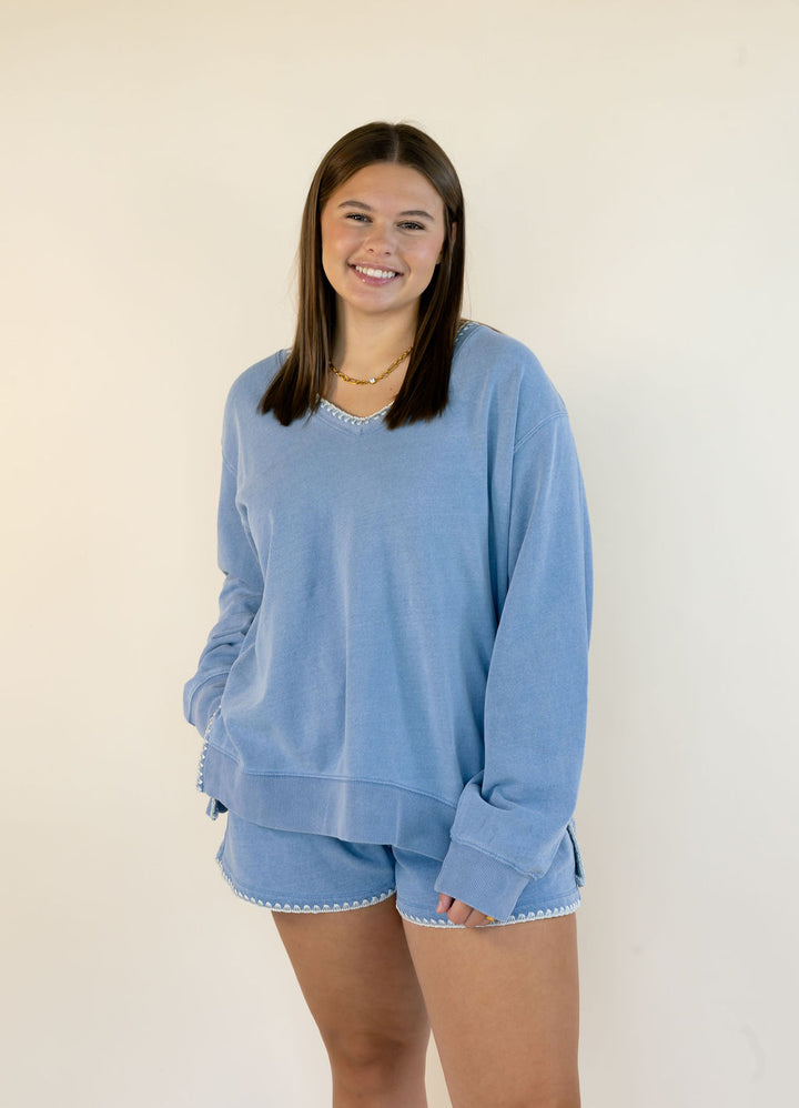 A smiling young woman wearing a light blue long-sleeve sweatshirt and matching shorts with white scalloped trim, standing against a plain off-white background.