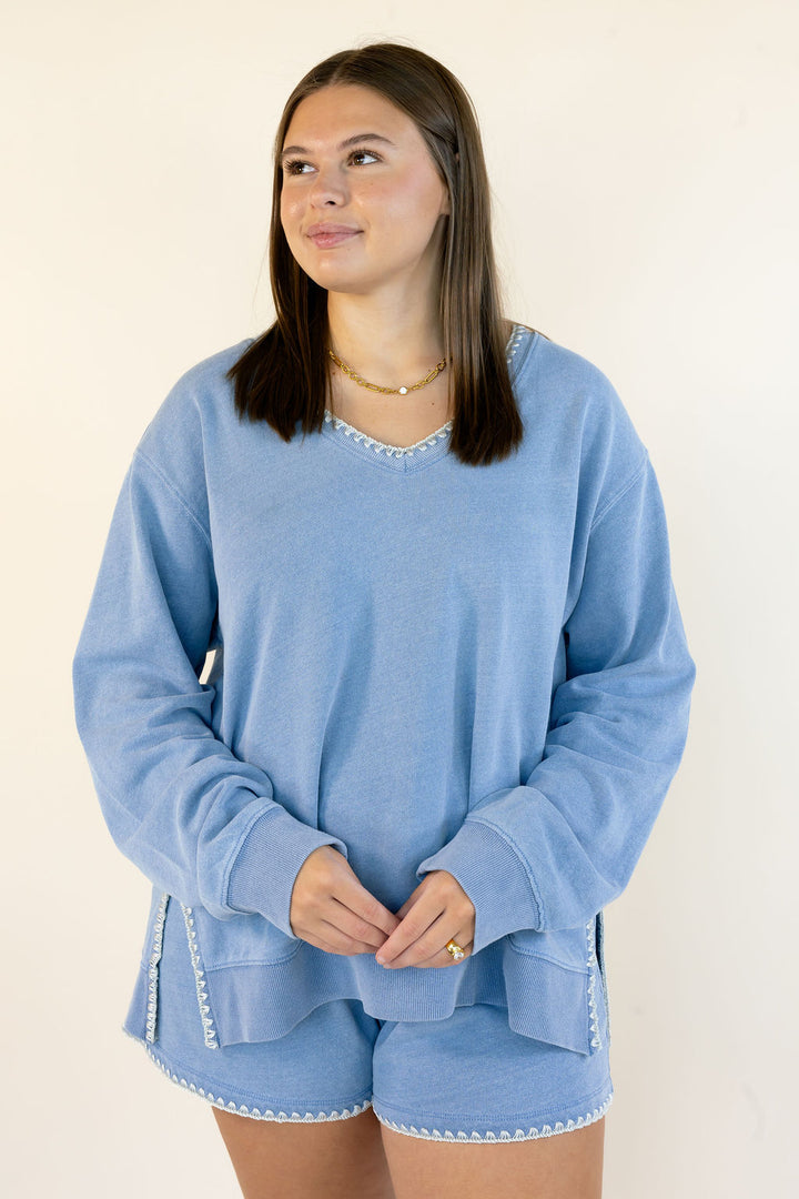A woman modeling a light blue lounge set consisting of a long-sleeve V-neck sweatshirt and matching shorts, both featuring white embroidered trim details, set against a plain off-white background.