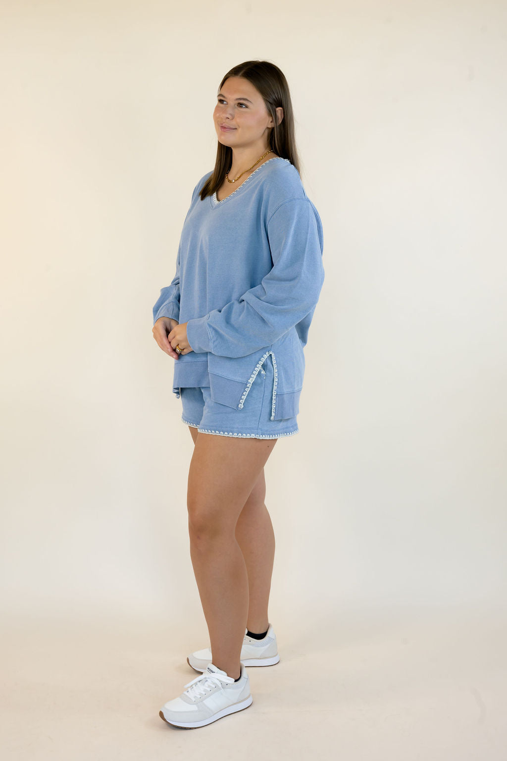 A woman modeling a light blue lounge set consisting of a long-sleeve V-neck sweatshirt and matching shorts with white trim, paired with white sneakers, against a plain cream studio background.