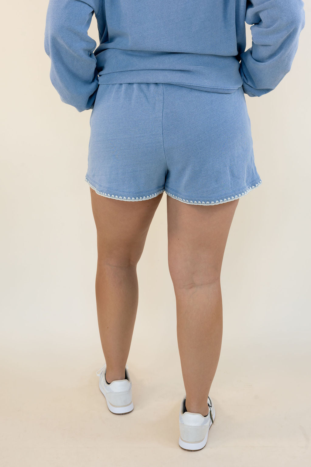 A person shown from the back wearing a light blue sweatshirt and matching shorts with white sneakers, against a plain beige studio background.