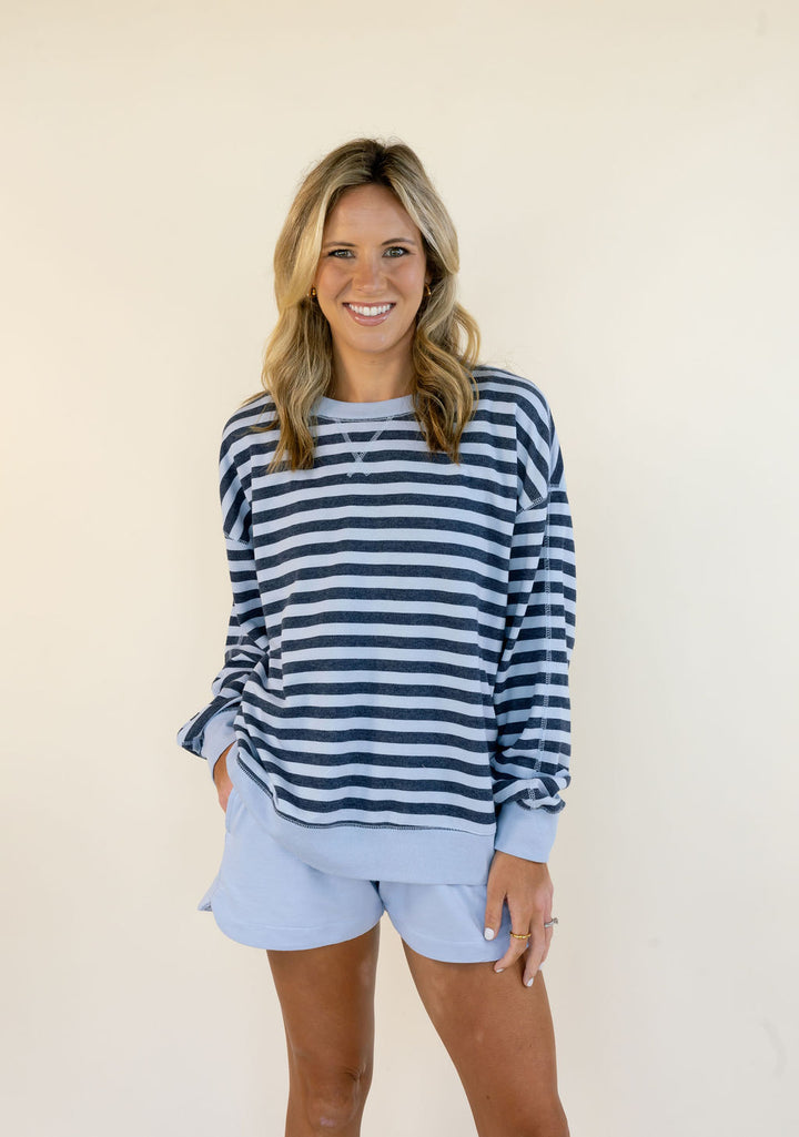 A smiling woman with blonde hair stands against a plain cream background wearing a navy and light blue horizontally striped long-sleeve top with matching light blue shorts.